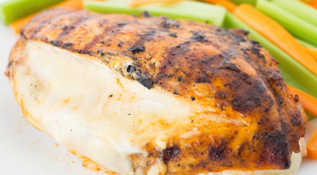 Close up of a grilled chicken breast that's been sliced down the middle and has white cheese oozing out of it. There's celery and carrot sticks out of focus in the background.