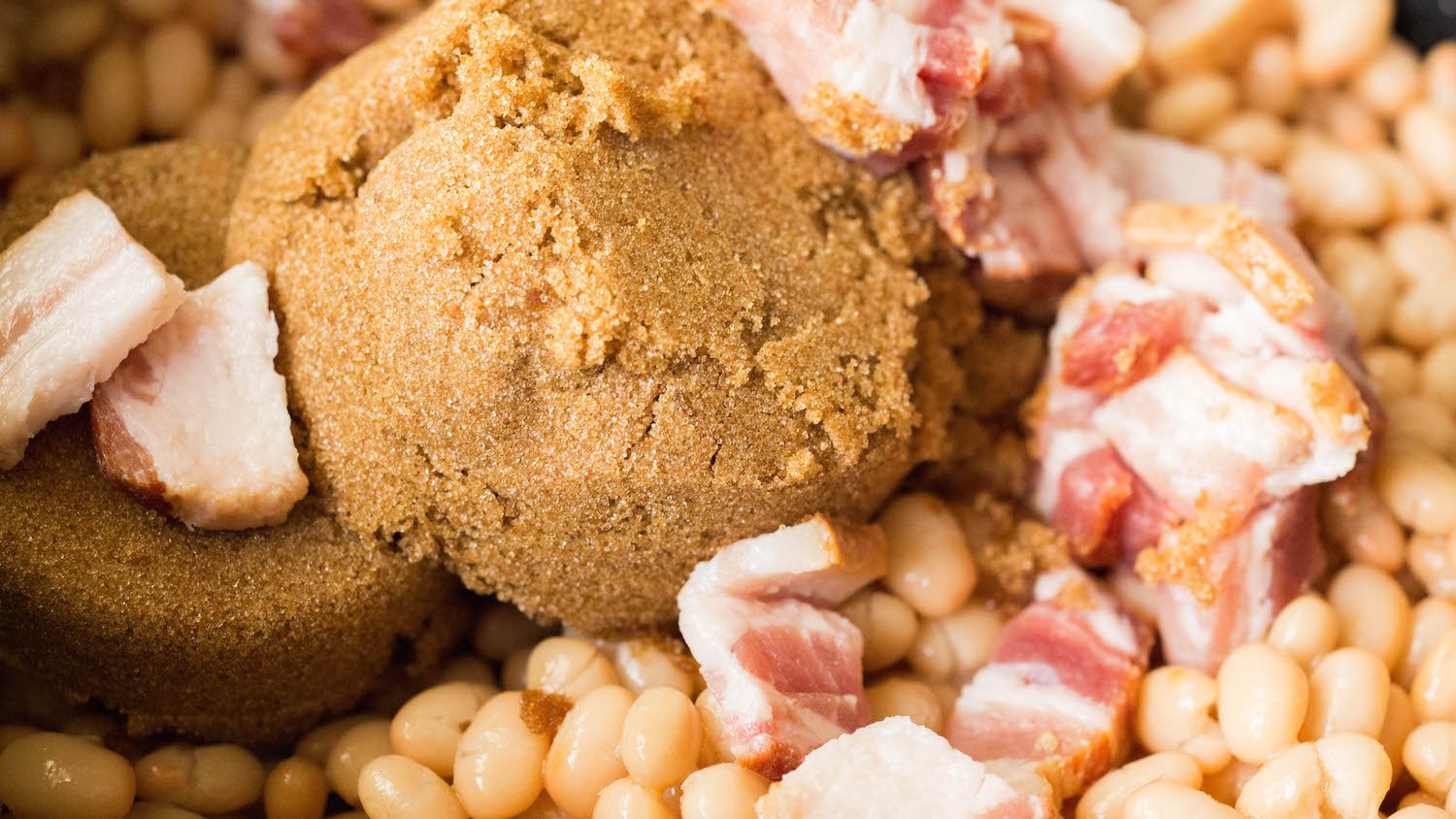 Close up of white beans, sliced bacon, and brown sugar in a pot.