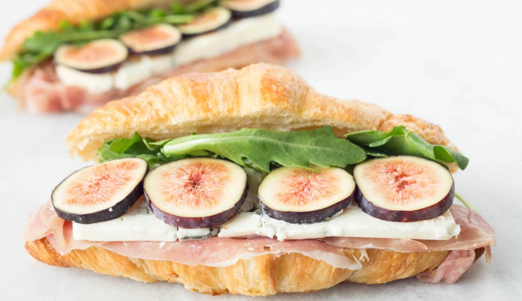 A croissant sandwich featuring prosciutto, blue cheese, sliced figs and arugula sits on a white background. Another fig sandwich is out of focus behind it.