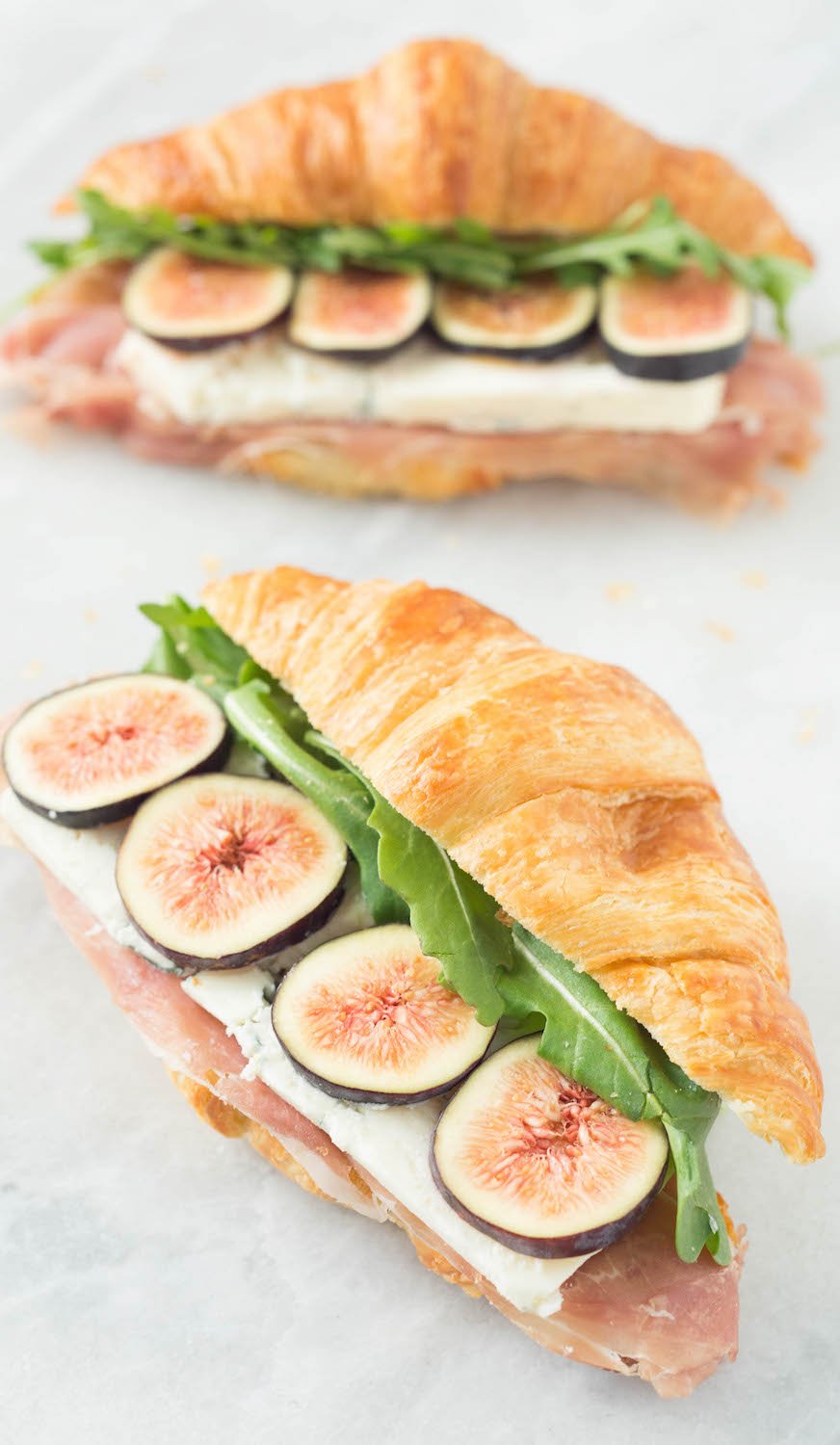 Two fig and blue cheese sandwiches sit on a white background. The croissant tops are positioned slightly off to the side so you can see the layers of prosciutto, blue cheese, sliced figs, and arugula. 