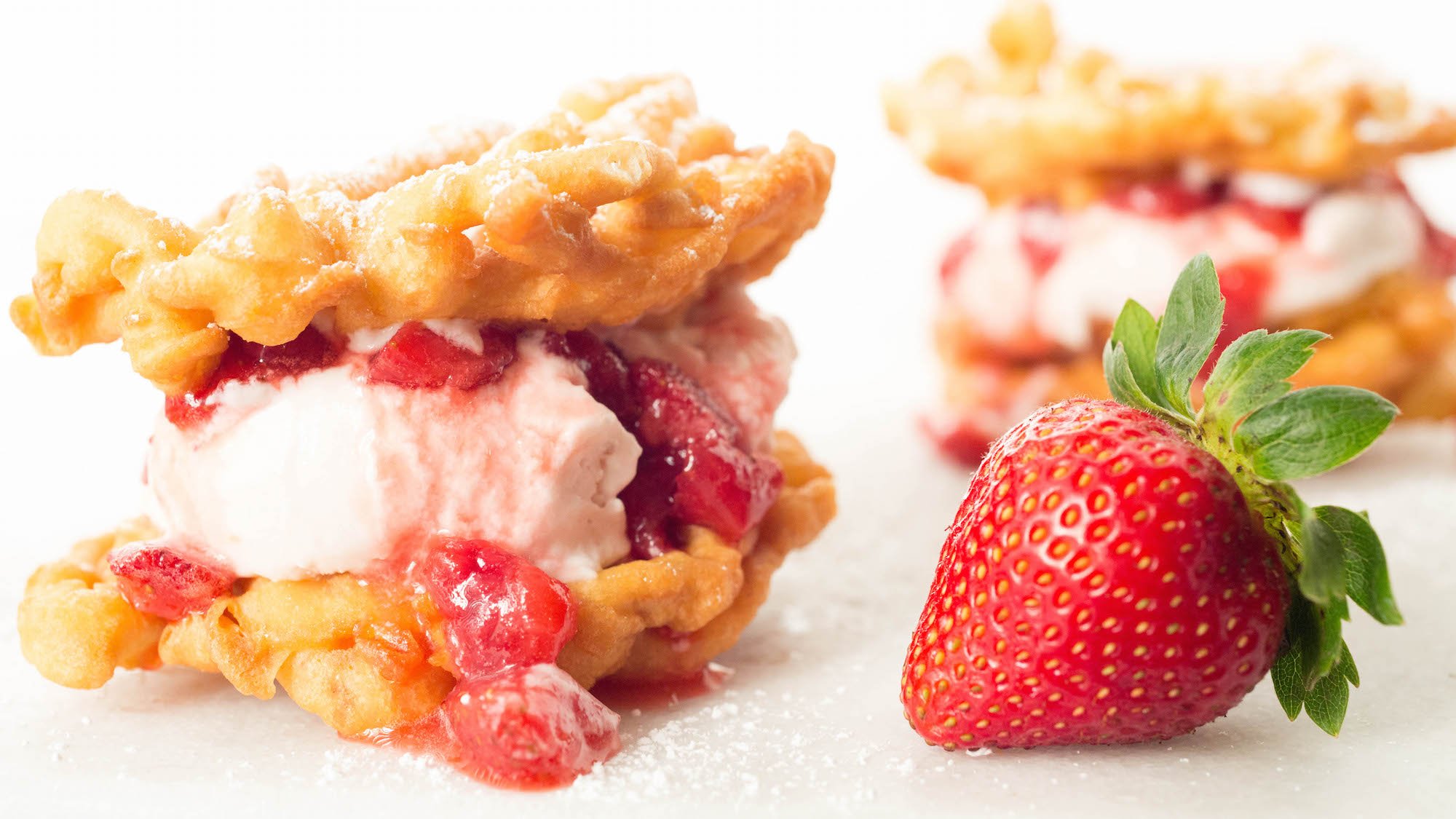 Two homemade Funnel Cake Ice Cream Sandwiches with Roasted Strawberry Buttermilk Ice Cream and fresh Strawberry Syrup on a white background next to a whole fresh strawberry