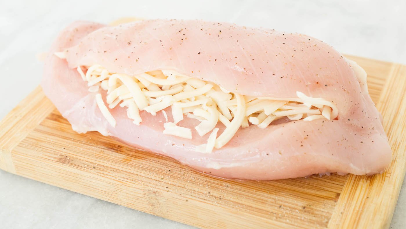 A raw chicken breast on a cutting board. It has been seasoned with salt and pepper, then sliced down the middle and stuffed with shredded mozzarella cheese.