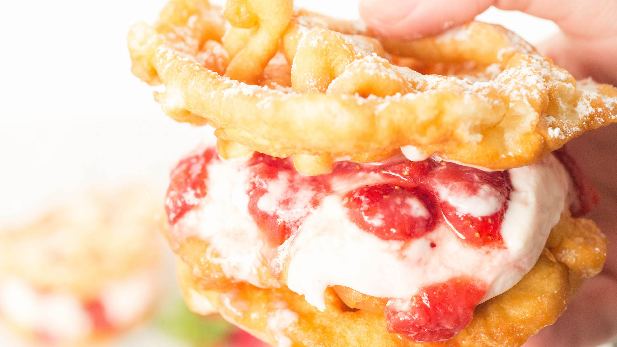 A hand holds a homemade funnel cake ice cream sandwich with strawberry syrup. 