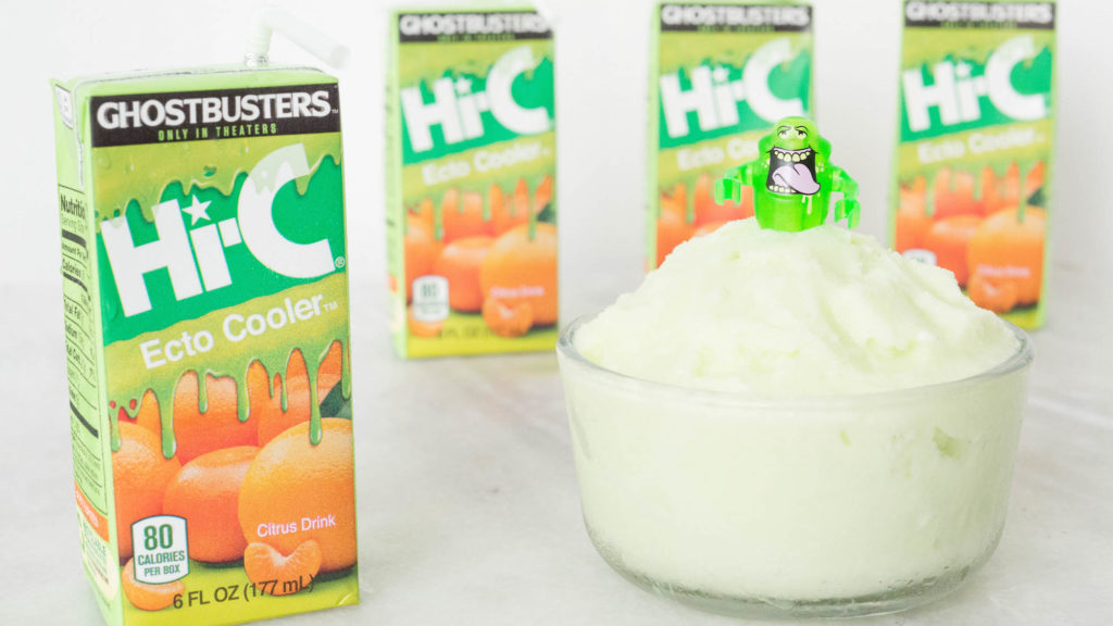 A small glass bowl has been filled with a light bright green colored sherbet. A lego version of the "Slimer" ghost from Ghostbusters sit on top. Cartons of Hi-C Ecto Cooler are arranged around the bowl.