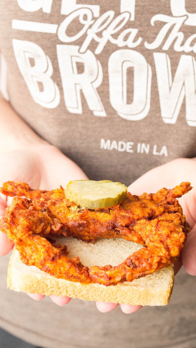 Two hands are holding up a slice of white bread that has an orange Nashville Hot soft shell crab that's topped with a pickle.