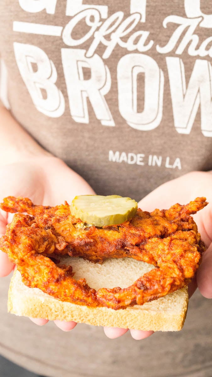 Two hands hold a slice of white bread with an orange Nashville Hot soft shell crab topped with a pickle.