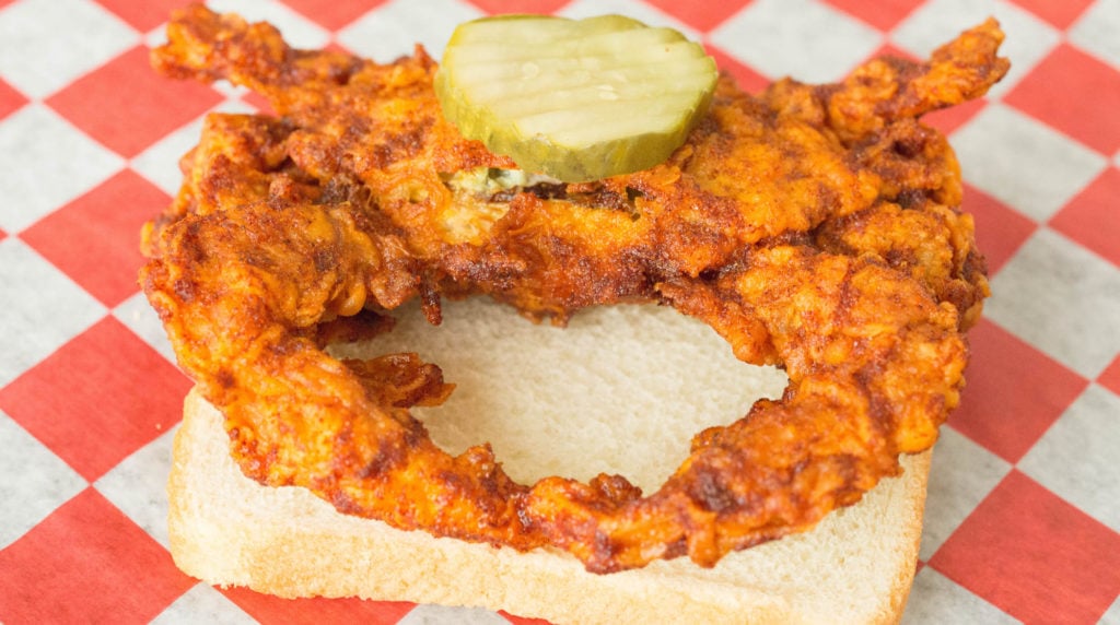 A soft shell crab that's been breaded, deep fried, and coated in Nashville hot sauce sits on a piece of white bread. There is a single pickle on top.