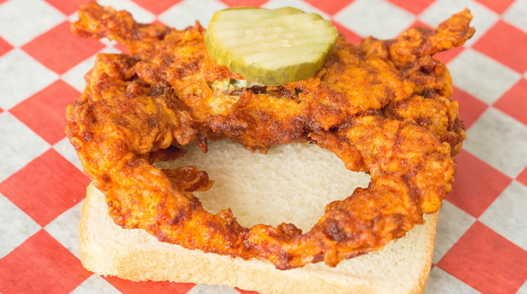 A soft-shell crab breaded, fried and covered in Nashville hot sauce sits on a piece of white bread.  There's only one pickle on it.