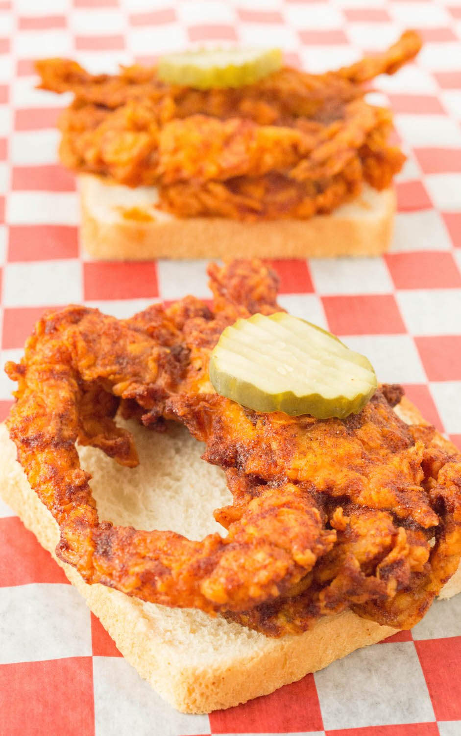 A deep fried soft shell crab that's been covered in Nashville hot sauce. It's sitting on top of a slice of white bread and has a pickle on top. Another one can be seen in the background.