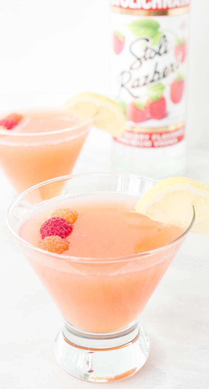 Two raspberry pink lemonade cocktails in stemless martini glasses sit on a white background. A bottle of Stoli raspberry vodka is out of focus in the background.