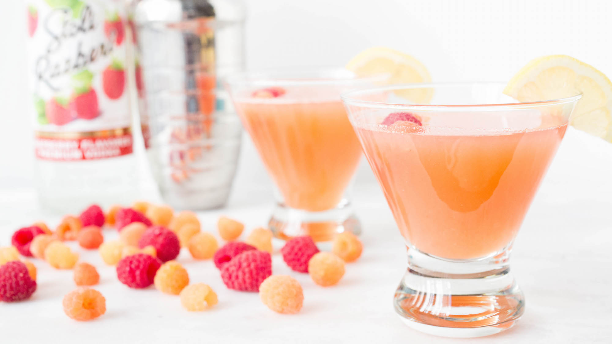 Two stemless martini glasses are on a white background. They contain pink lemonade raspberry cocktails. Pink and red raspberries are arranged around the glasses. A bottle of raspberry vodka and metal shaker are out of focus in the background.