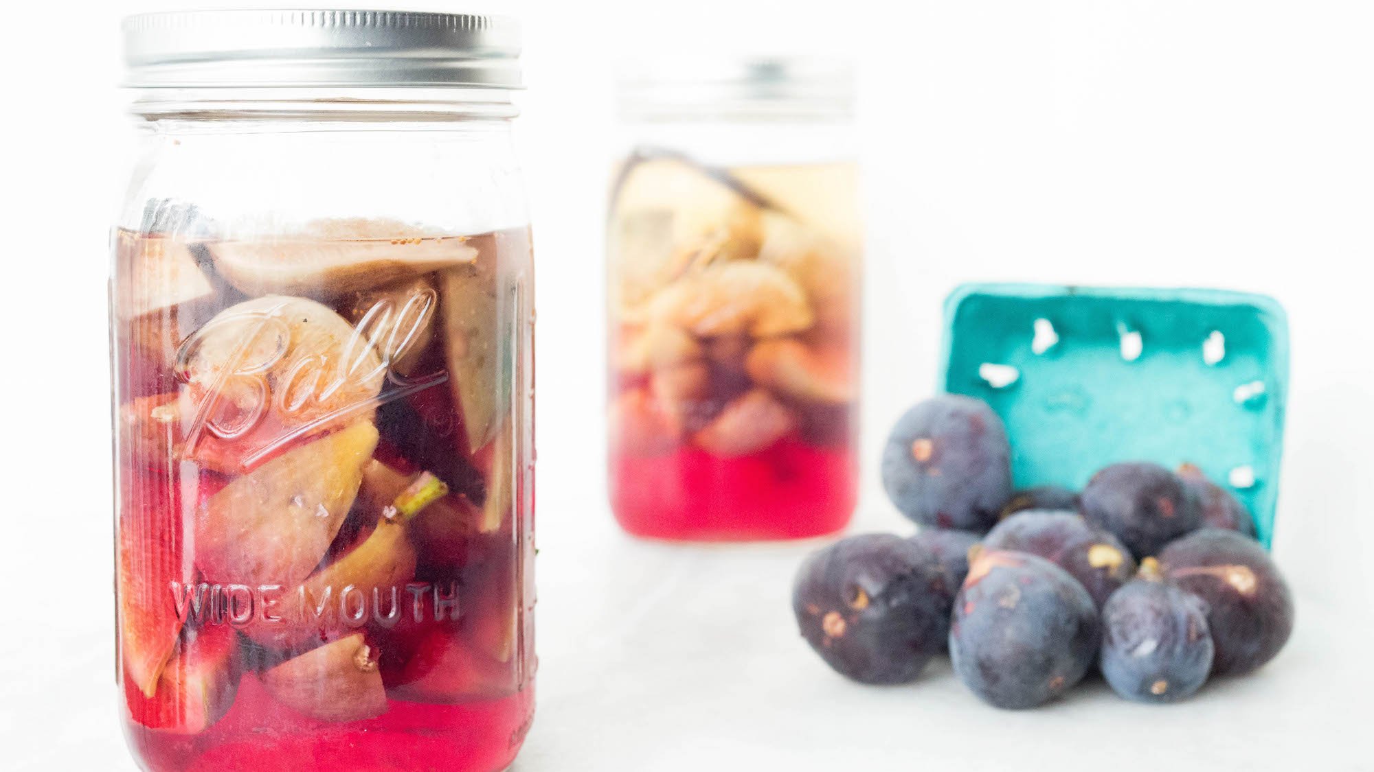 A large glass mason jar is filled with vodka that's turned pink from the sliced figs inside. Another jar is out of focus in the background along with a basket of fresh brown figs.