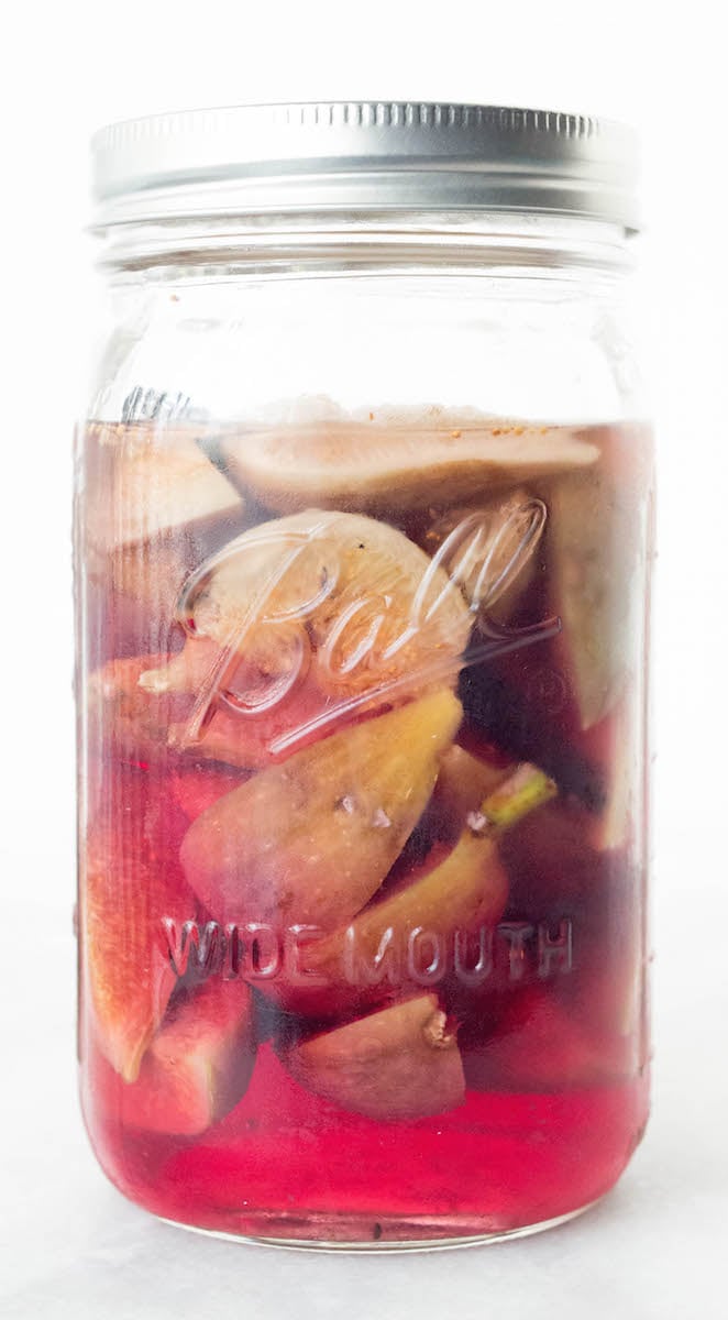 A jar of pink tinted homemade fig infused vodka siting on a white background.