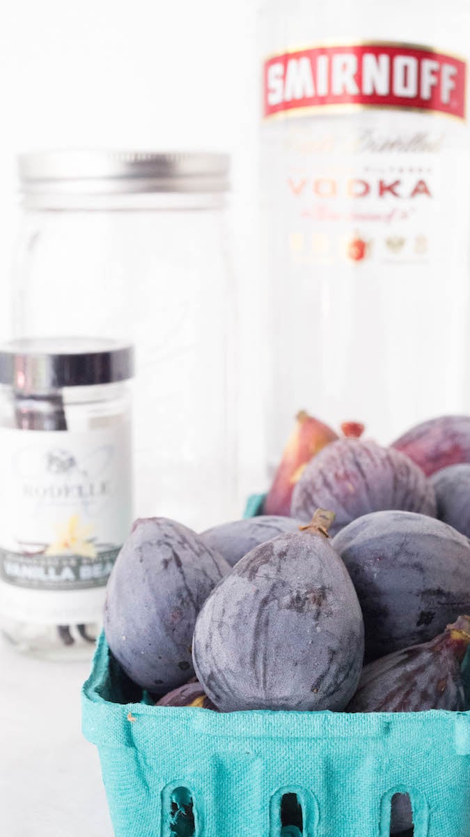 All the ingredients for fig infused vodka sit on a white background. A small basket of fresh brown figs, a bottle of Smirnoff vodka, a jar of vanilla beans, and an empty mason jar.