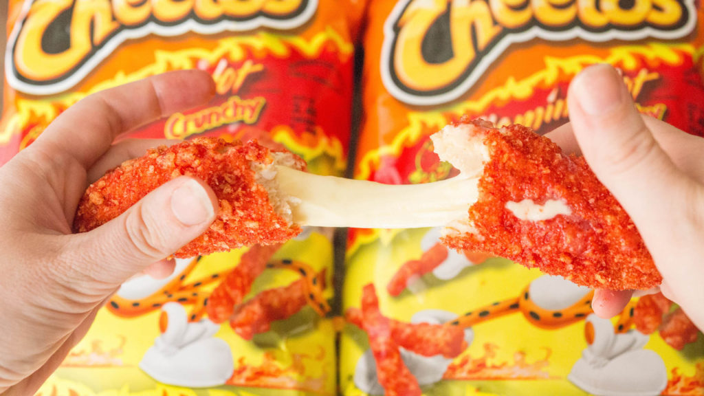 A cheese stick covered in bright orange flamin' hot Cheetos coating is being pulled apart to reveal the melted cheese over two bags of flamin' hot Cheetos.