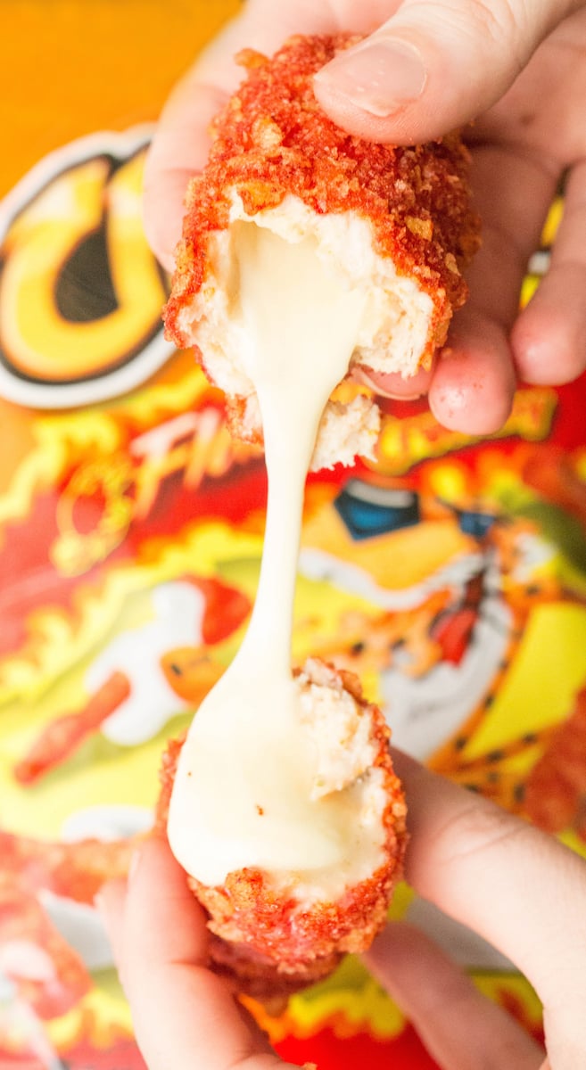 A close up of a mozzarella cheese stick coated in flamin' hot Cheetos dust being pulled apart by two hands showing the melted cheese scretch.