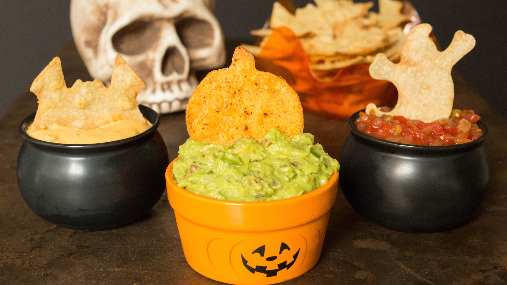 All three Halloween tortilla chips in dips In front of a skull. 