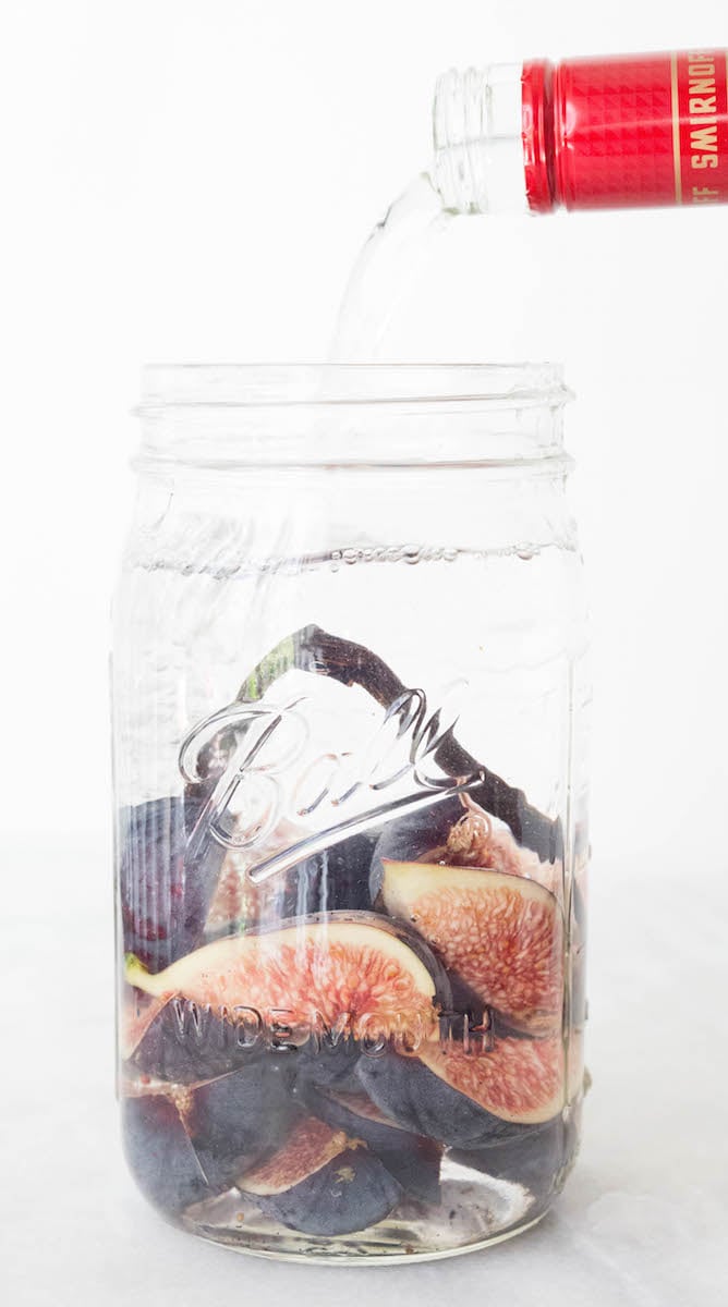 A large mason jar has fresh sliced figs and a vanilla bean inside of it. Vodka is being poured over them.