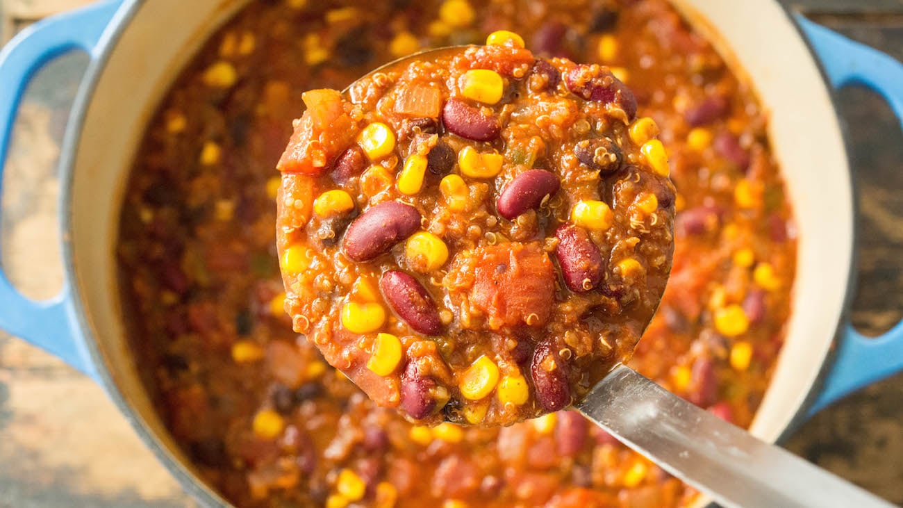 Overhead a dutch oven for that's filled with meatless chili. A metal ladle holds up a scoop of it so you can see the individual beans, tomato, corn, and quinoa.