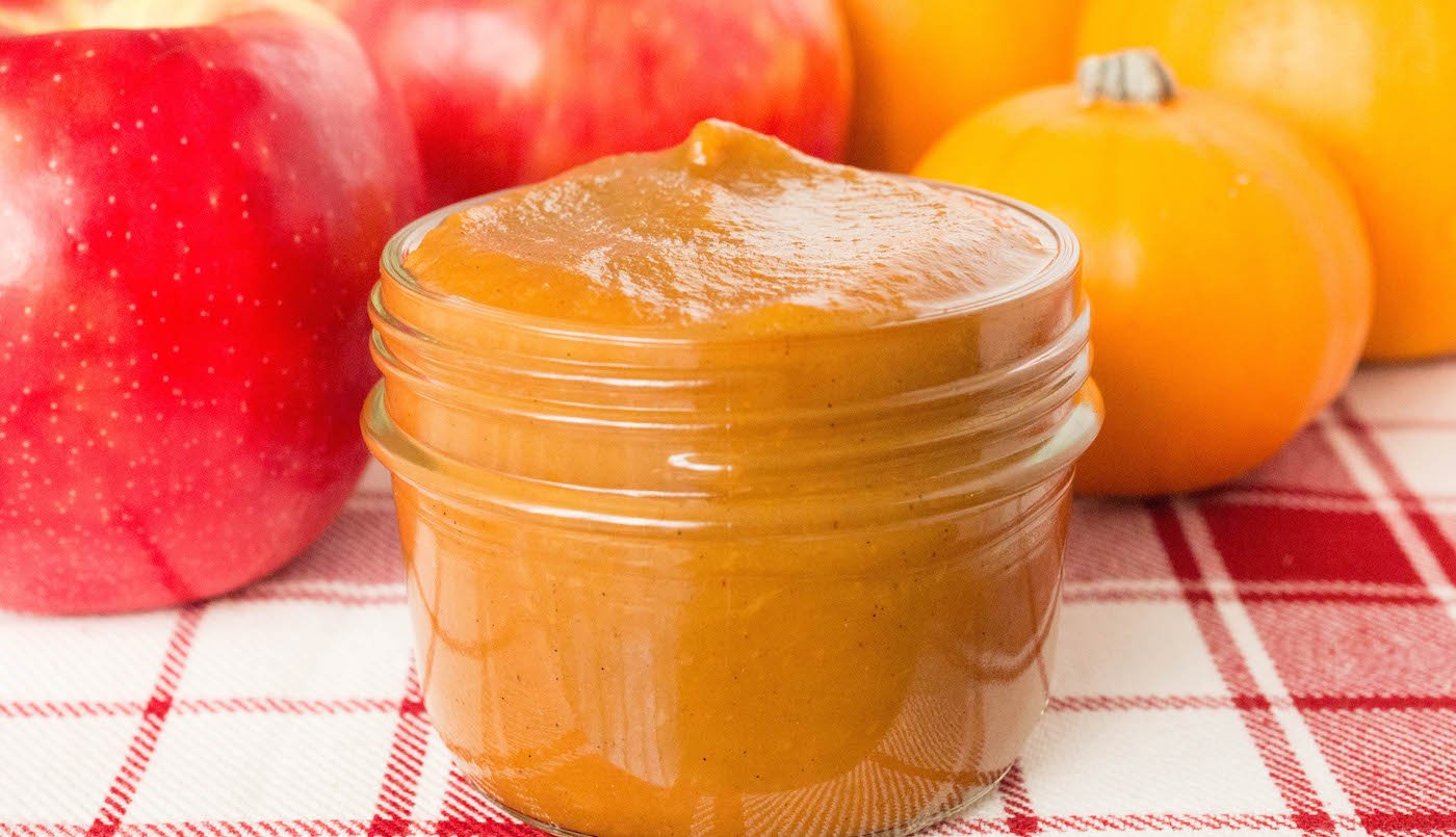 A small glass mason jar filled with pumpkin apple butter is in focus in the foreground, small pumpkins and a few apples are out of focus in the background.