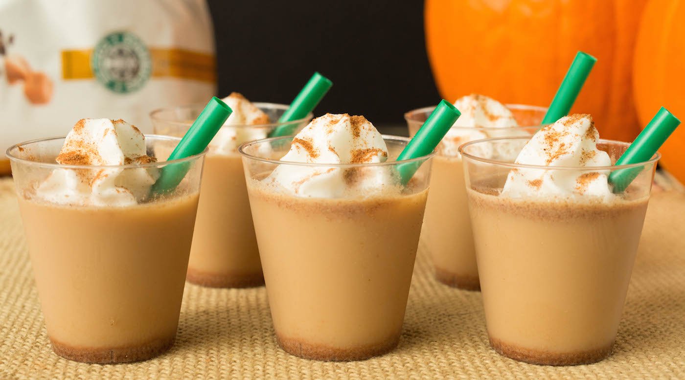 5 pumpkin spice latte jello shots in mini see through plastic cups have short green straws in them. Out of focus in the background is a Starbucks coffee bag and an orange pumpkin.