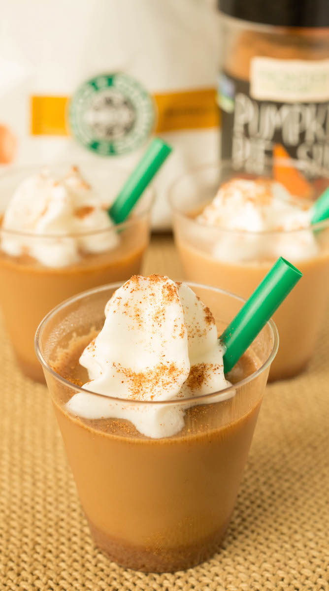 Close up of a pumpkin spice latte in mini see through plastic cup with a dollop of whipped cream on top and a green plastic straw on the side.