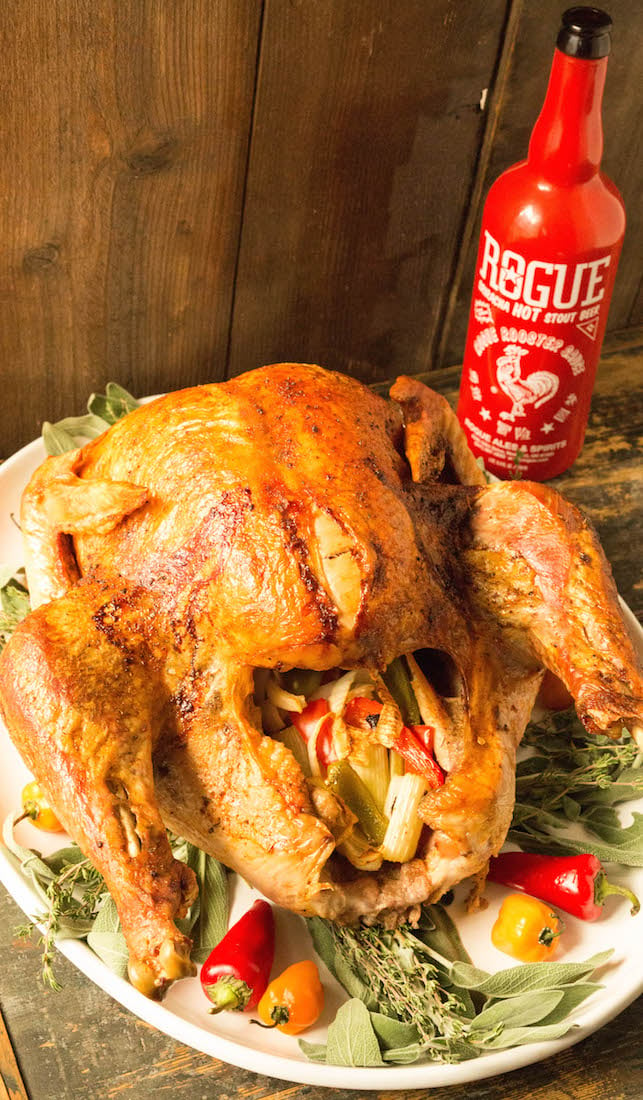 A large cooked cajun turkey sits on a white platter in front of a bottle of Sriracha beer. The platter is garnished with fresh herbs and spicy peppers. They turkey's cavity is filled with chopped bell peppers, celery, and onion.