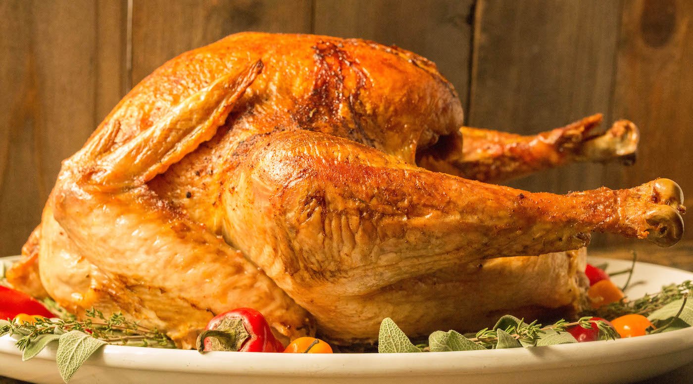 A beautifully browned cooked cajun Turkey sits on a large white platter. It's garnished with fresh herbs and fresh habanero and Fresno peppers.