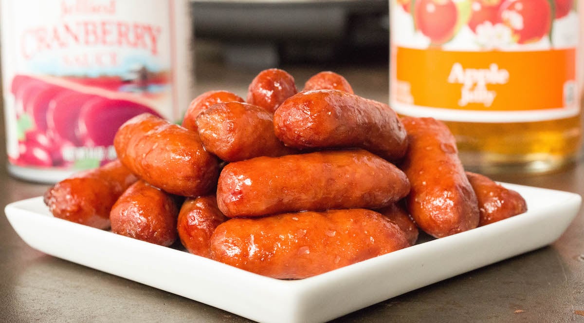 Apple Cranberry lil' smokie sausages stacked on a small white plate in front of apple jelly and canned cranberry sauce.