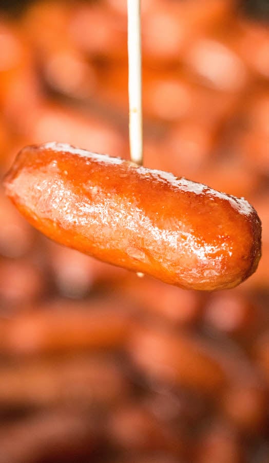 Close up of a toothpick holding a single apple cranberry lil smokie in front a crock pot full of them.