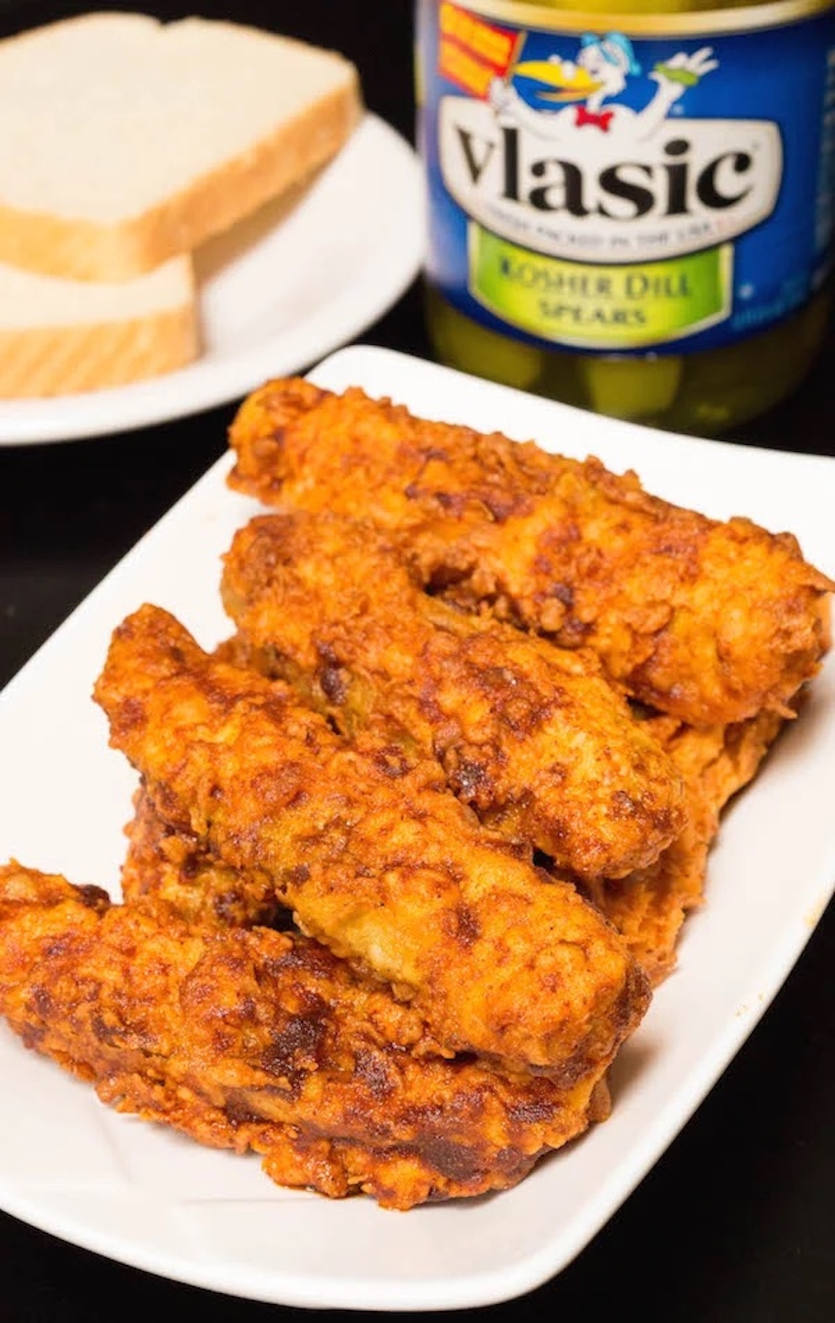 Breaded fried pickle spears coated in a bright orange sauce in a small white serving dish. A jar of pickles and slices of white bread are out of focus in the background.