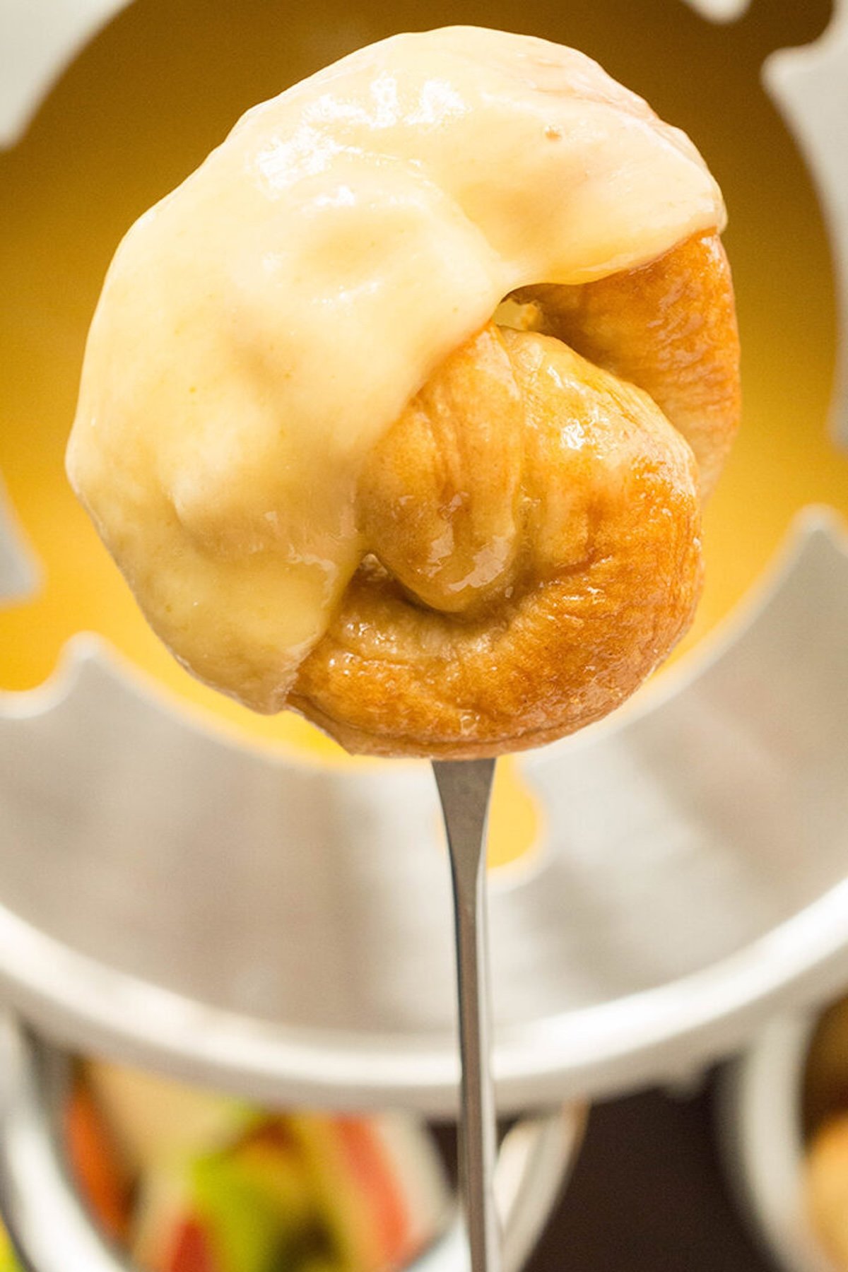 A mini soft pretzel that's been dipped in yellow fondue beer cheese is on a fondue skewer, the pot of fondue cheese is out of focus in the background.
