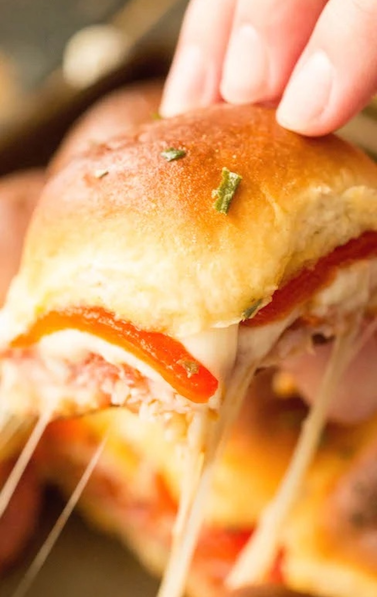 A hand pulls a single Hawaiian roll slider filled with Italian meats and red peppers out of a pan to show the melted cheese pulling.