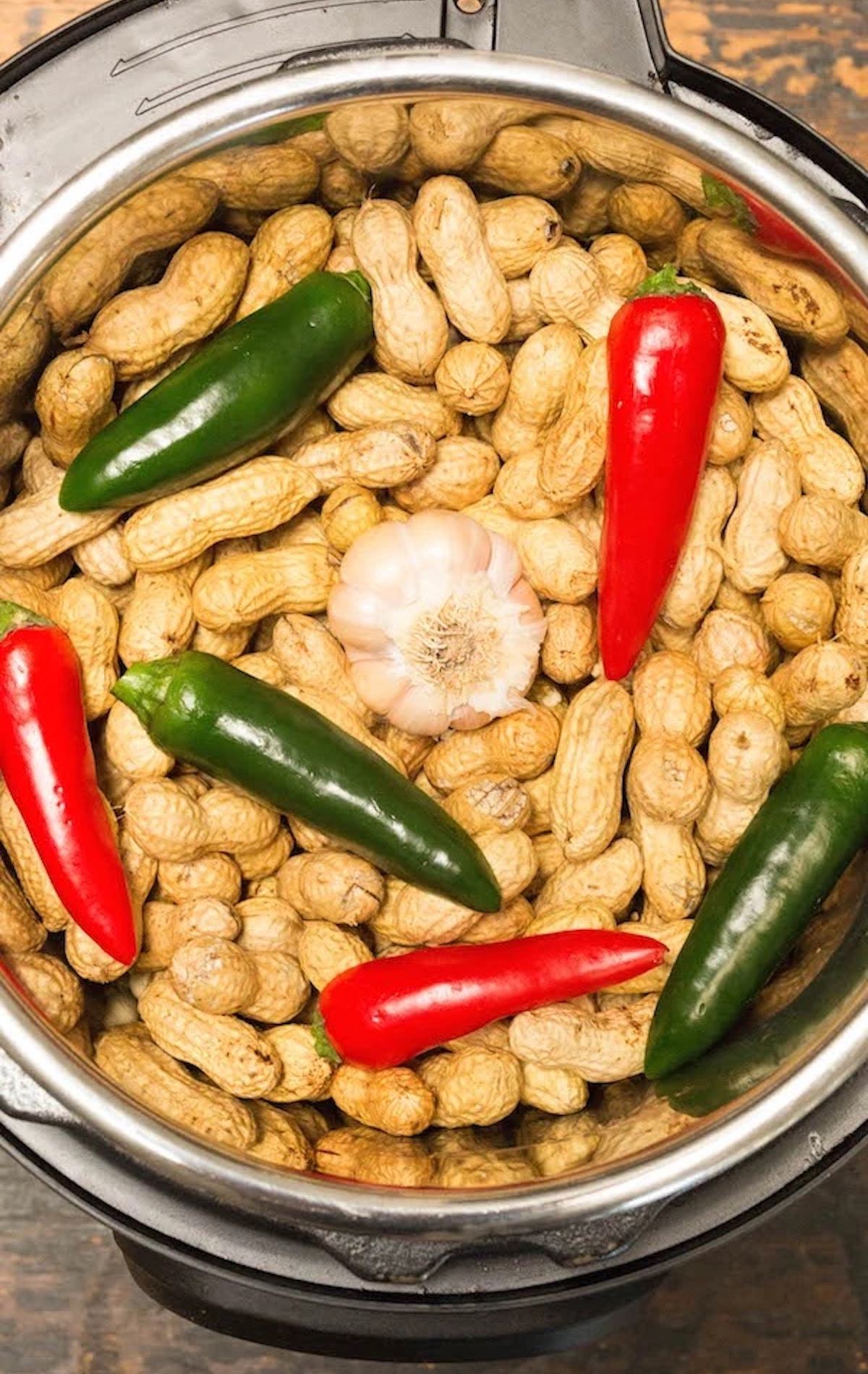 Overhead view of an Instant Pot filled with peanuts in their shell, several whole green and red peppers, and a whole bulb of garlic.