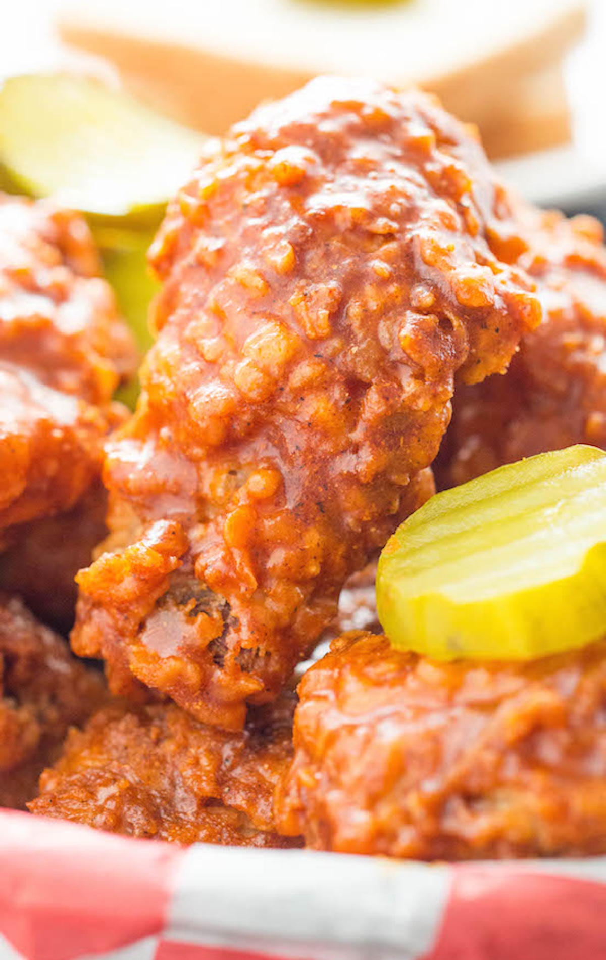 A breaded fried chicken drummette that's coated in a reddish orange sauce with visible flecks of spices. More wings and dill pickle chips are out of focus in the background.