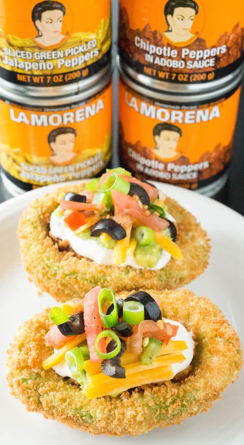 Two deep fried seven layer stuffed avocados in front of cans of La Morena Chipotle Peppers and Jalapeños. 