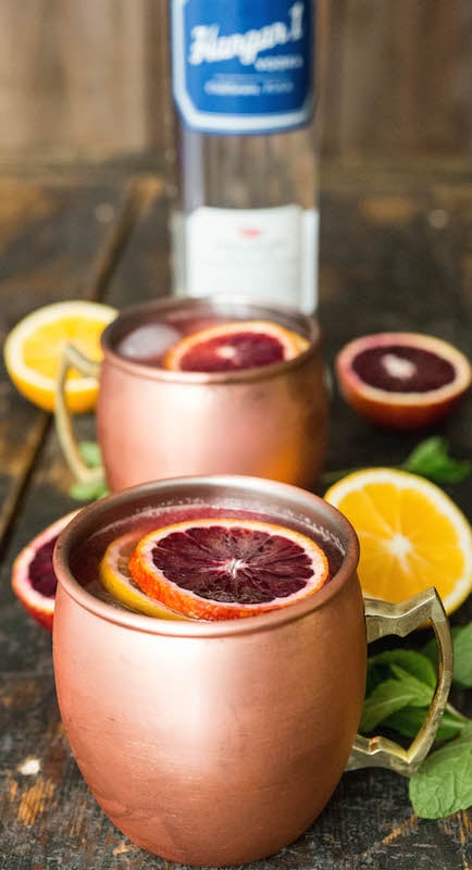 A copper mug filled with blood orange & Meyer lemon Moscow Mule is garnished with a slice of each citrus. Another Moscow Mule and a bottle of vodka are out of focus in the background.