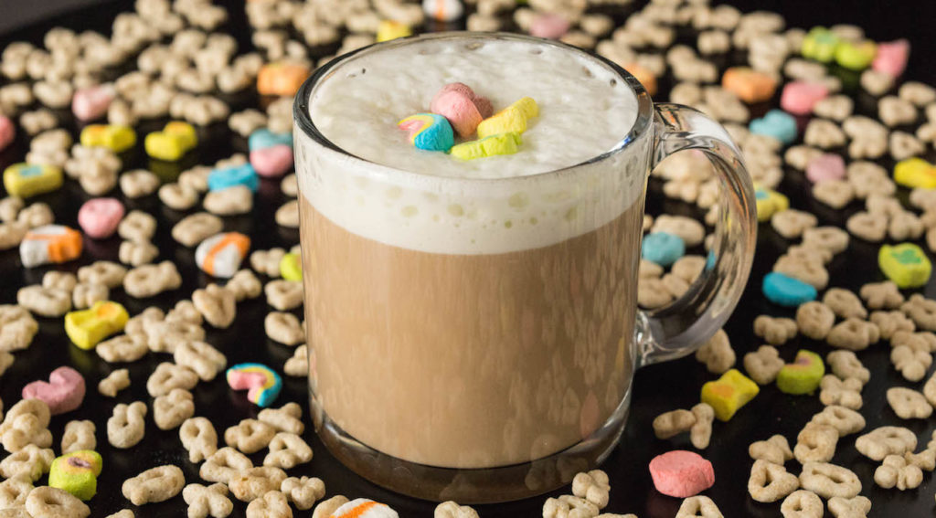Lucky Charms Cereal Milk Latte in a clear coffee mug surrounded by the cereal. 