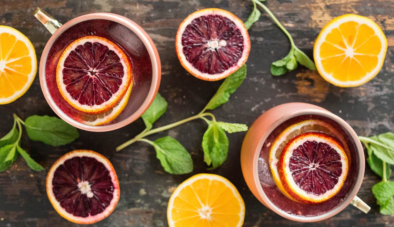 Overhead view of two copper mugs filled with blood orange and Meyer lemon Moscow Mule. Mint sprigs and sliced citrus are arranged around the cups.