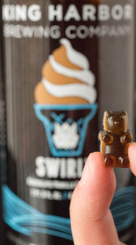 Close up of fingers holding a single beer gummy bear with a beer bottle out of focus in the background.