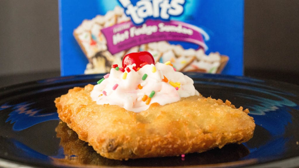 A deep fried pop tart has been topped with whipped cream, sprinkles, and a cherry. It sits in front of an our of focus Pop-Tart box.
