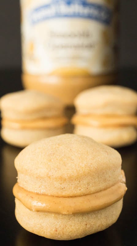 A close up of a peanut butter filled macaroon dog treat. More treats and a jar of peanut butter is out of focus in the background.