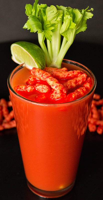 A Flamin' Hot Cheetos Bloody Mary on a black background. It's garnished with some of the Cheetos, a lime slice, and a celery stalk. 