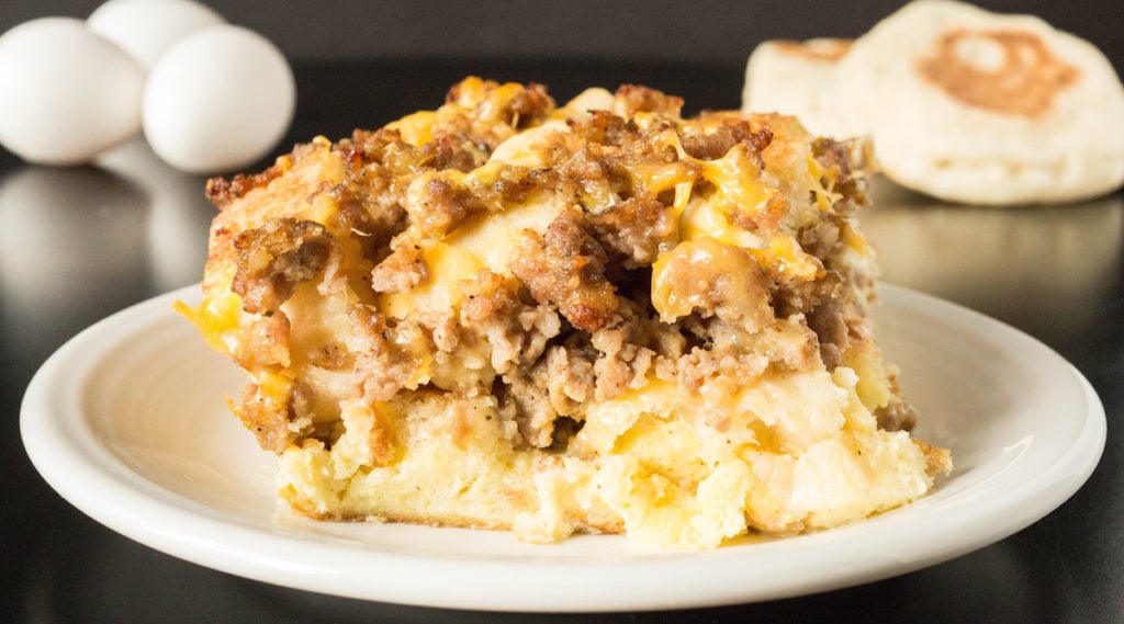 A slice of Sausage Egg McMuffin Casserole with eggs and English muffins out of focus in the background.