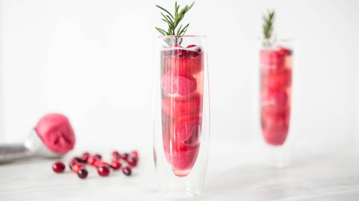 Two clear champagne flutes filled with Cranberry Sorbet Rosé Mimosas. A scoop of the sorbet is out of focus along with some fresh cranberries in the background.
