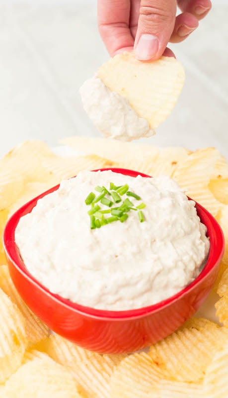 A hand hold up a potato chip that's been dipped in Instant Pot Caramelized Onion dip over the bowl of it.