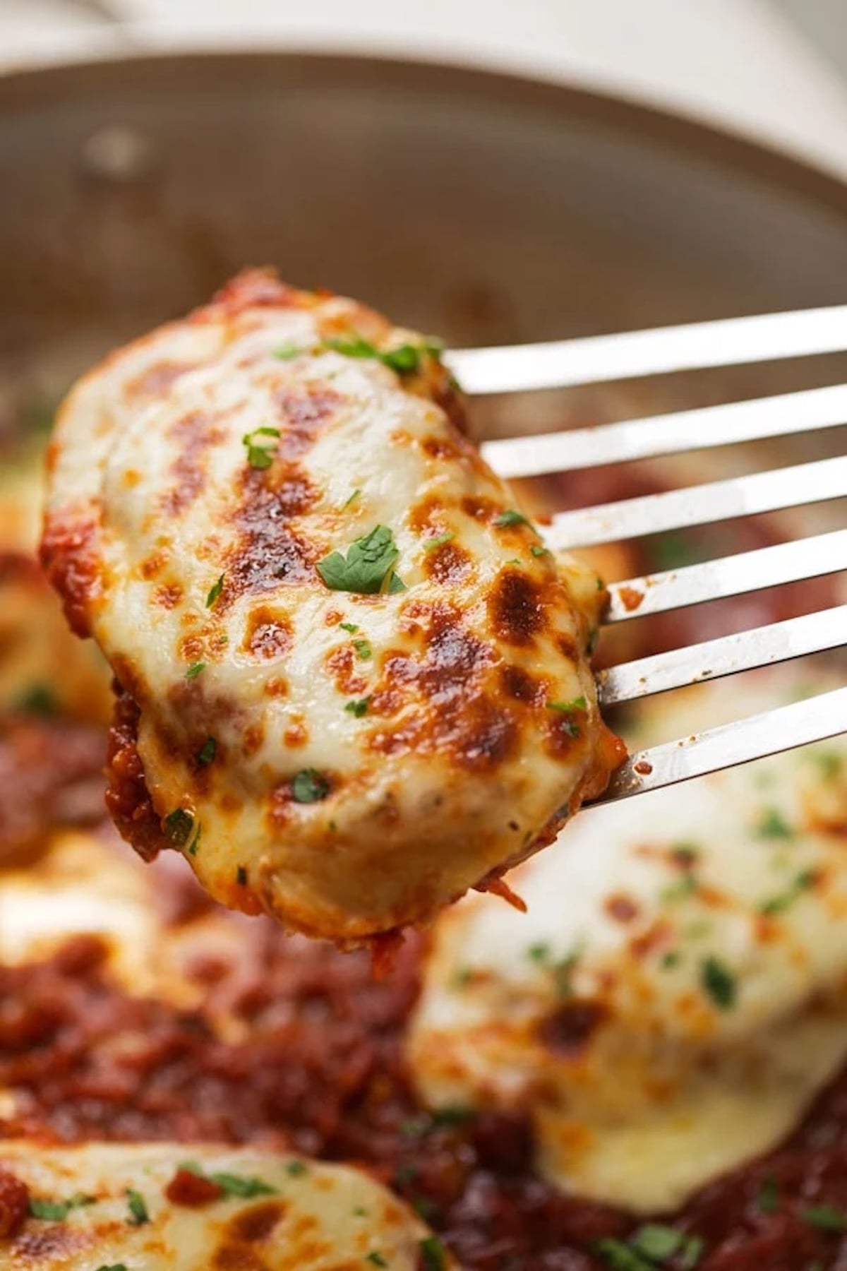 A spatula lifts a chicken breast that is completely coated in melted, browned mozzarella cheese. A skillet with the rest of the cheese covered chicken is out of focus in the background.
