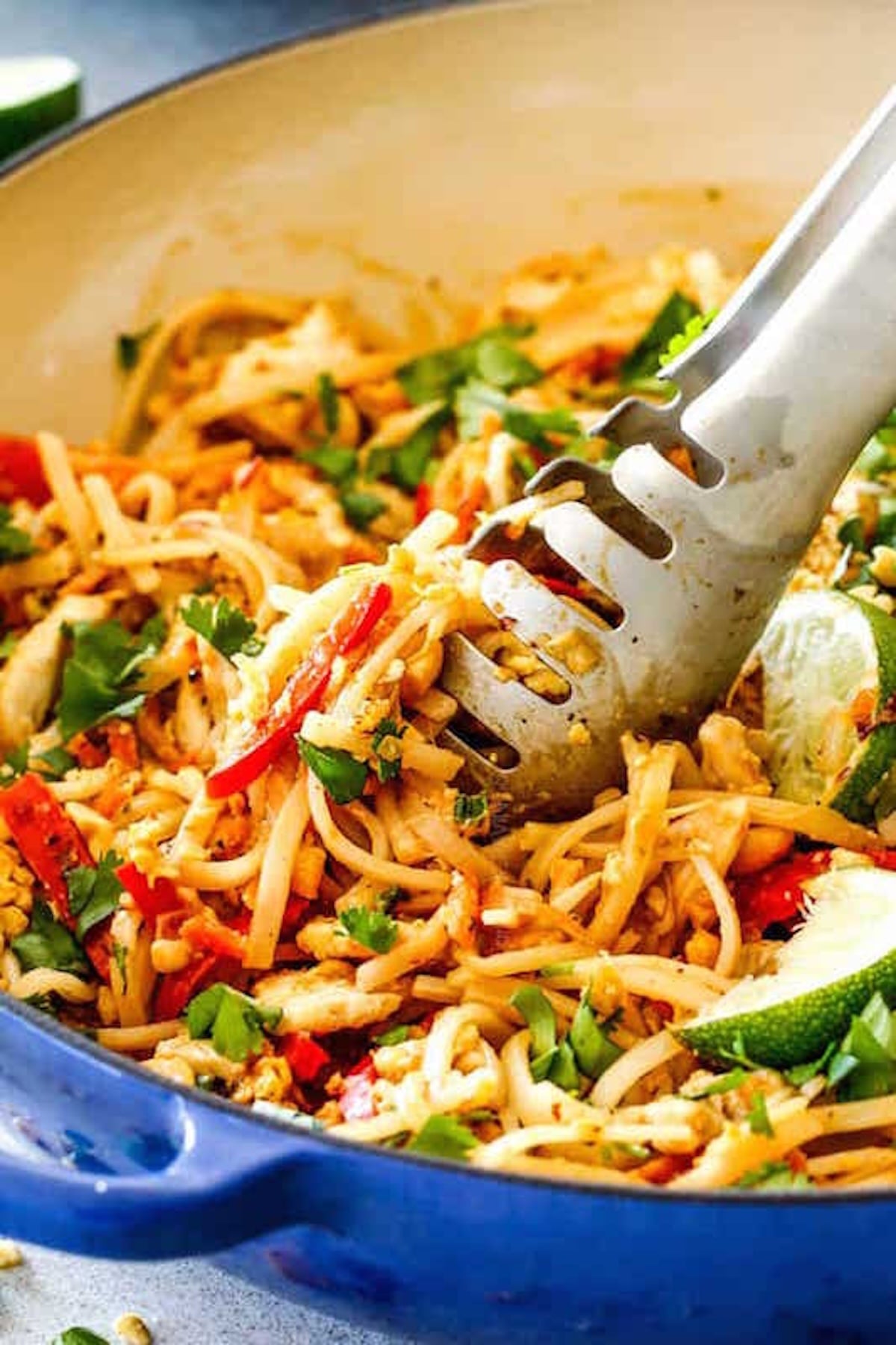 A large skillet filled with a chicken pad Thai that as visible chopped herbs, red peppers, and lime slices.