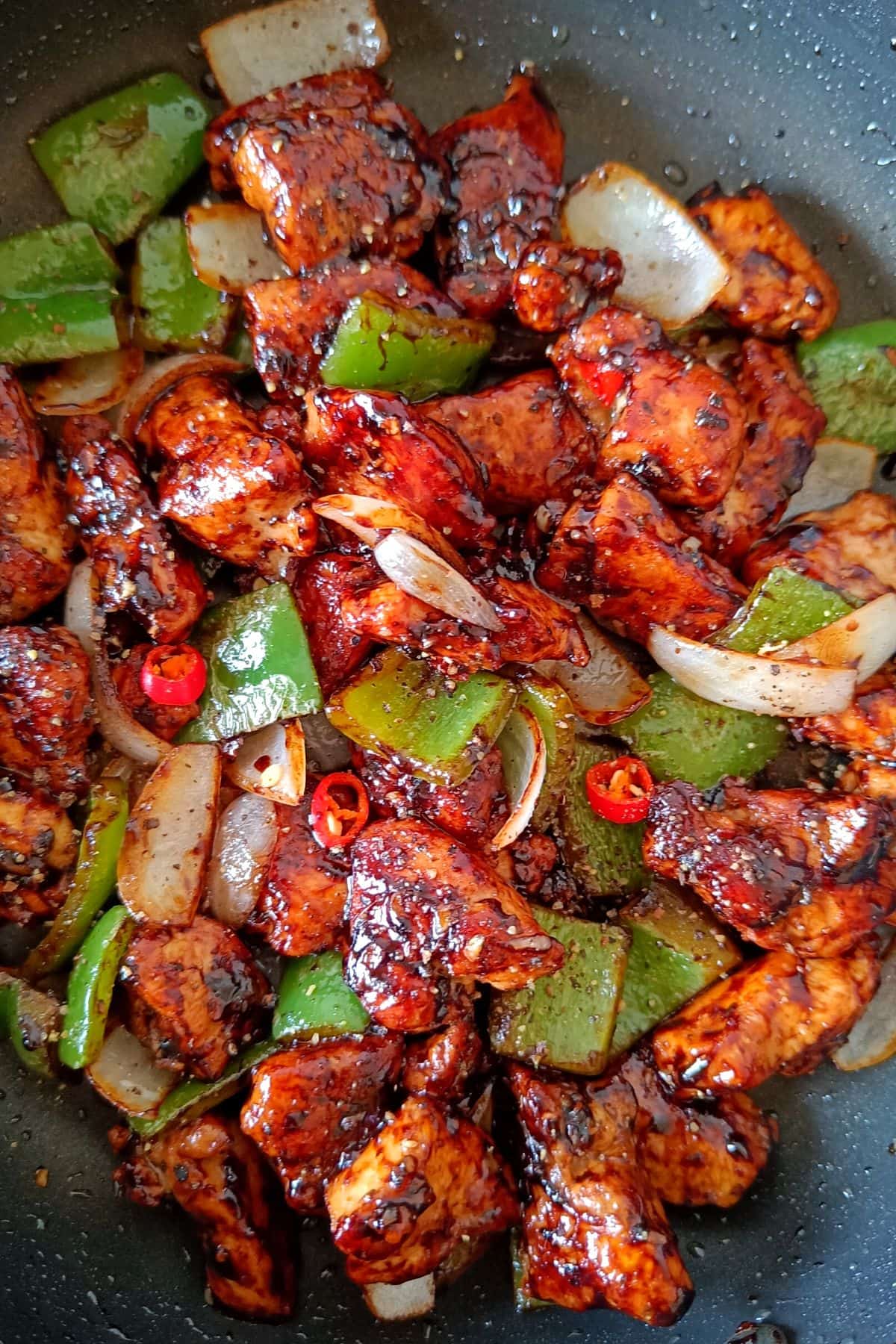 Chunks of chicken coated in a deep red glaze in a skillet with largely diced green bell peppers, white onion, and red hot peppers.