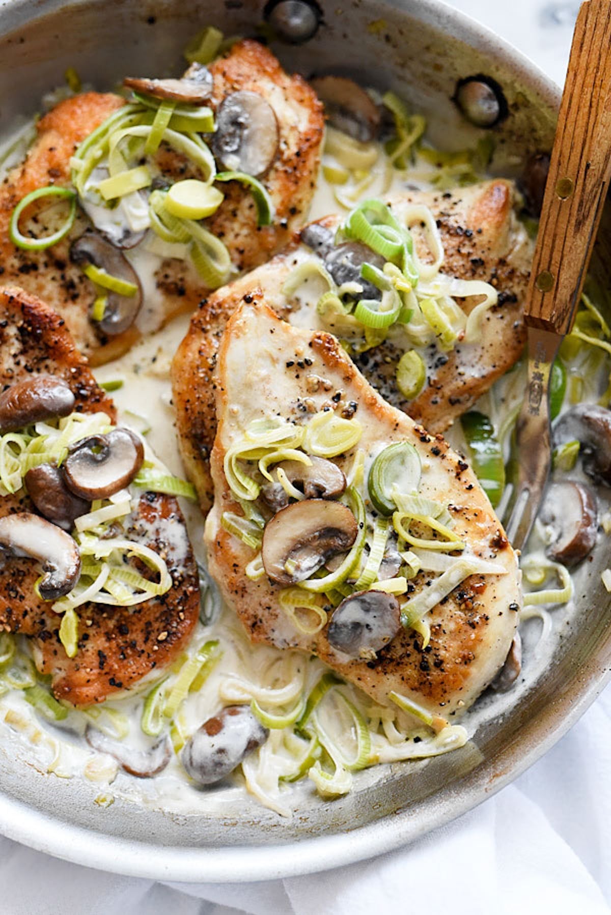 A stainless steel skillet is filled with seared chicken breasts that are topped with sliced mushrooms and leeks. They are sitting in a creamy white sauce with additional mushrooms and leeks.